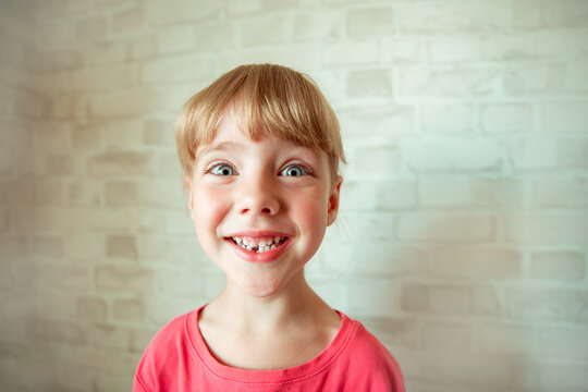 joyful girl with big expressive eyes and a wide happy smile, with a short bob haircut with bangs, in a pink T-shirt, against the background of a white brick wall shows a tooth that has fallen out