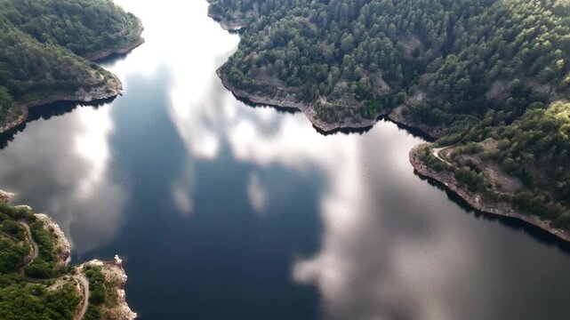 Lake and Cloud Reflection. Aerial view of a winding mountain lake, reflecting bright white fluffy clouds like a mirror, surrounded by dense green slopes of coniferous forest.