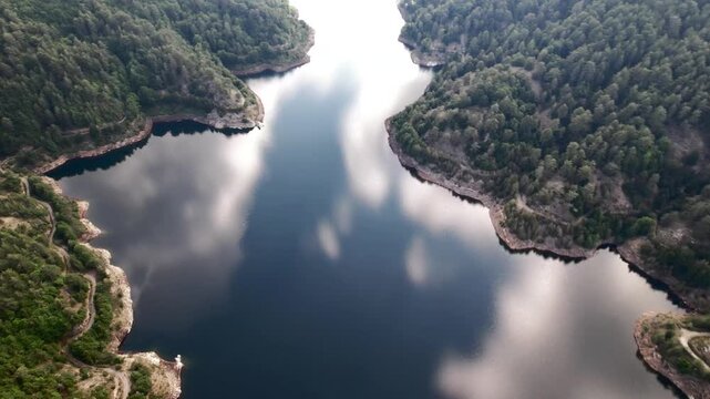 An aerial view of a blue lake reflecting clouds. The lake, meandering through steep hills covered in dense green forest, reflects the detailed contours of the shoreline and clouds in its waters.