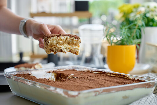Tiramisu slice held above glass dish, showing cocoa topping and creamy layers on a kitchen counter