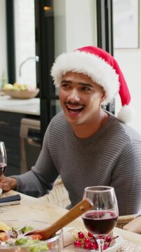 Vertical video: Laughing man in Santa hat hearing friend speaking, turning at table with wine glass