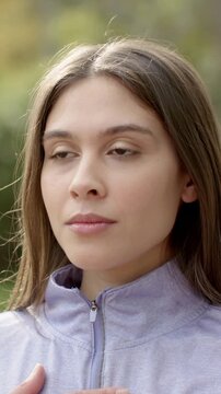 Vertical video: Opening eyelids woman in light purple zip-up jacket smiling in park, hand on chest