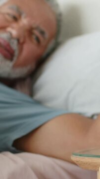 Vertical video: Feeling pain, man in blue shirt reaching arm toward bedside pillow and glass table