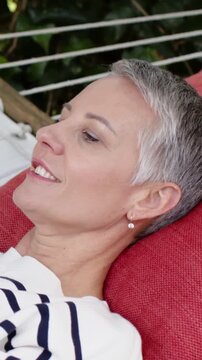 Vertical video: Seeing offscreen, woman reclining on hammock in garden laughing in striped sweater