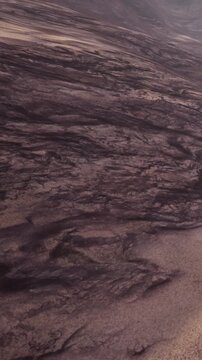 A dry lava field shows black rock formations with textures and patterns. The area is mostly barren with shades of tan soil visible. The landscape appears rugged and uneven.