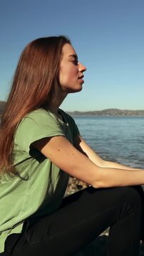 Vertical video: Sitting lady green tee tilting head to bay, sun causing calm on rocks, copy space