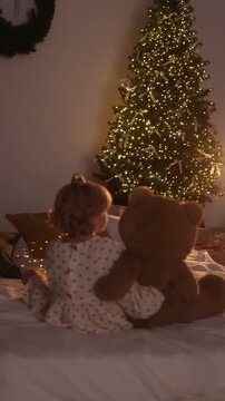 Young toddler sits on bed with arm around oversized brown teddy bear, turning head toward tall evergreen tree decorated with warm white lights, creating soft glowing bokeh in cozy bedroom