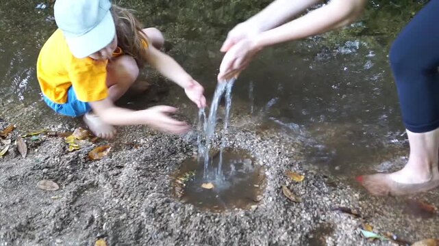 Close-up of a mother pouring water from her cupped hands into a small sand pool built by her child barefoot in a shallow forest stream.