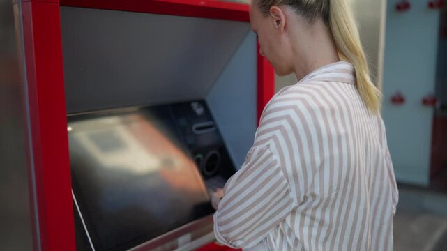 Young blonde woman interacting with an automated teller machine on street, carefully entering personal identification number and using touchscreen to complete transaction