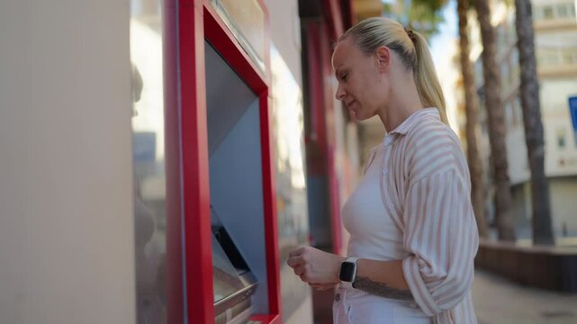 Blonde woman stand on city street withdrawing cash from an atm, inserting card, enter pin, and retrieving money from banking machine for personal finance
