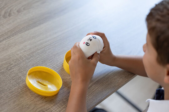 Young child grips white squishy toy with printed cartoon face beside open yellow capsule on wood grain tabletop, with small hands, soft texture, indoor daylight, and playful everyday detail