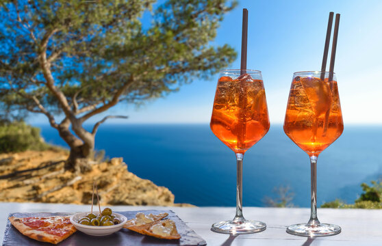 Two Aperol Spritz are on a sunny terrace next to snacks and olives overlooking the blue Mediterranean Sea &ndash; a relaxed summer coastal atmosphere.