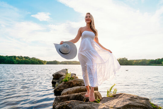Woman in white dress stands on rocks by lake during sunny day with hat in hand