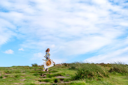 Woman walking on a green hillside with a straw hat and a large tote bag during a bright day