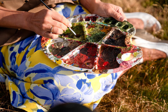 Artist holds a paint palette and works on a landscape in a grassy field during daylight hours
