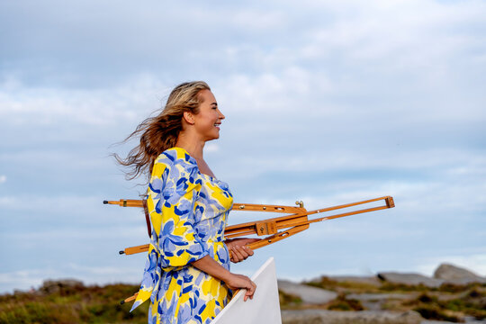 Artist walks along the beach carrying an easel and canvas under a cloudy sky