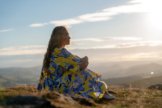 Person sits on a hilltop, enjoying the view during sunset with mountains and clouds in the background