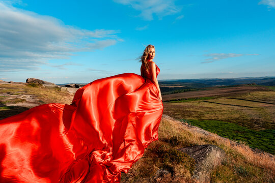 Woman in a red dress stands on a hilltop looking out over a valley during sunset with open sky and green fields in the background