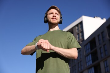 Fototapeta premium Man in sportswear with headphones checking fitness tracker near building under blue sky, low angle view