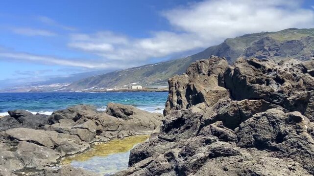 Rocky coastline and natural swimming pools with crystal blue ocean and mountain backdrop on a sunny summer day in Los Silos, Tenerife, Canary Islands, Spain, 4K.