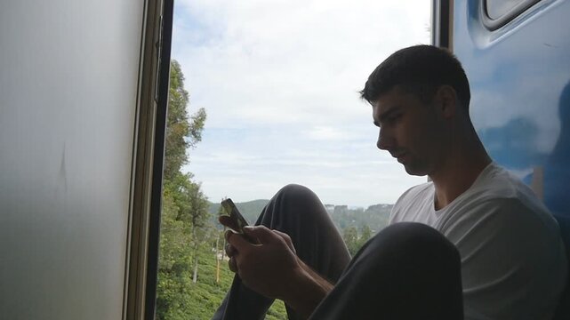 Male tourist sits near open door of moving train and uses smartphone. Young man rides on passenger railway transport and browses photos on mobile phone. Concept of summer travel or tourism. Slow mo