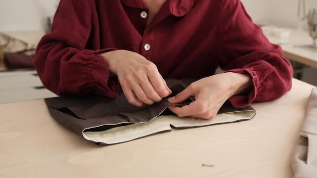 A tailor in linen shirt carefully hand-sewing the lining of dark trousers on a light wooden table inside a bright creative studio. Slow fashion, traditional tailoring, DIY sewing tutorials, craft work