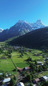 Vertical aerial shot over village houses in a valley, revealing a green mountain and the iconic rocky, snow-covered peak of Mount Ushba in summer.