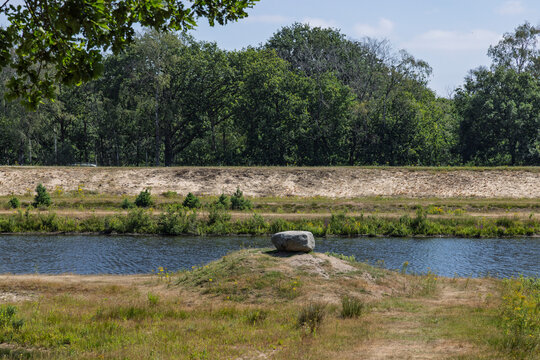 Hilversum, Netherlands. A large glacial erratic boulder sitting on a grassy mound next to a canal with a dense forest background in the nature reserve.