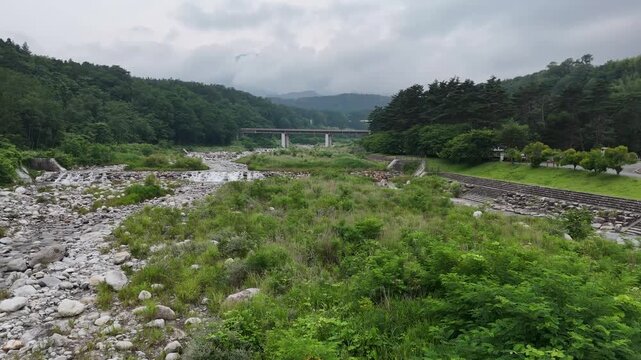 River flows gently through green, rocky banks. Clouds hang low above forested hills and a distant bridge. Grassy slopes border the stream, edged with stone retaining walls
