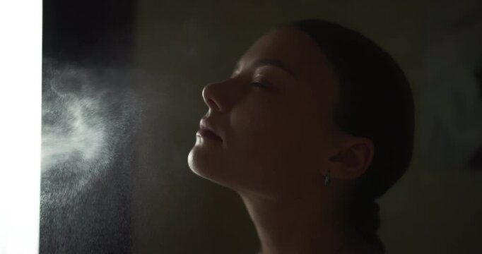 Beautiful girl refreshing skin with thermal water spray in dark room, slow motion