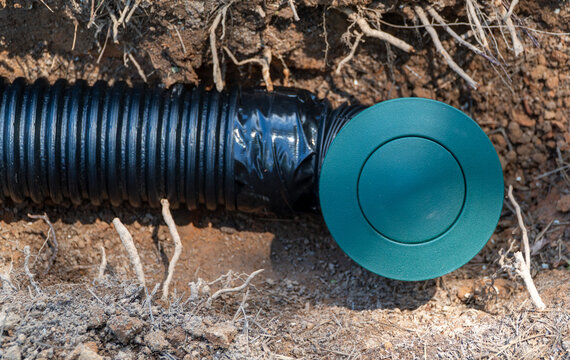 Underground black corrugated drainage pipe with a green end cap buried in dry soil, showing roots and dirt, for residential or landscape water management and erosion control.