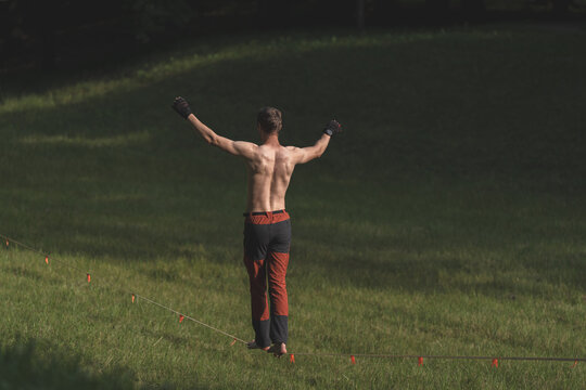 Shirtless man balancing on slackline with arms outstretched on grassy hillside, outdoor slacklining sport and balance training concept.