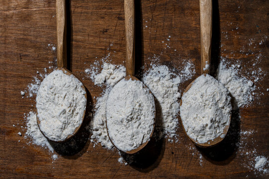 Three wooden spoons filled with white flour on rustic wooden background, closeup. Top view of white flour in traditional wooden spoons on dark rustic table background with spilled powder