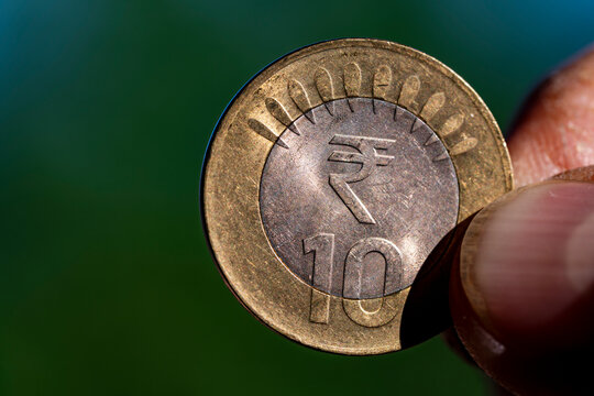 Closeup of hand holding Indian ten rupee coin on green background. Macro shot of person holding an official 10 Indian rupee bimetallic coin with blurry green nature background