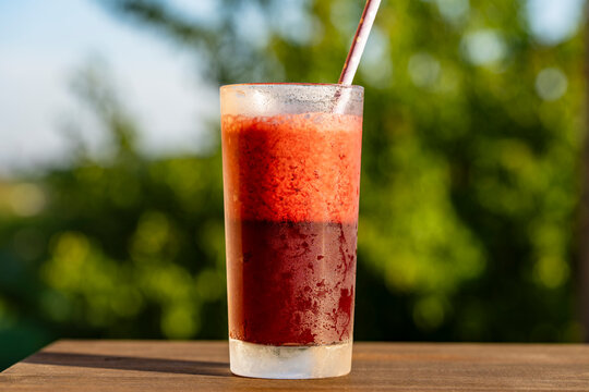 Fresh cold berry smoothie in glass with straw on blurred green background, closeup. Chilled red fruit smoothie with condensation drops and drinking straw on wooden table over blurry nature background