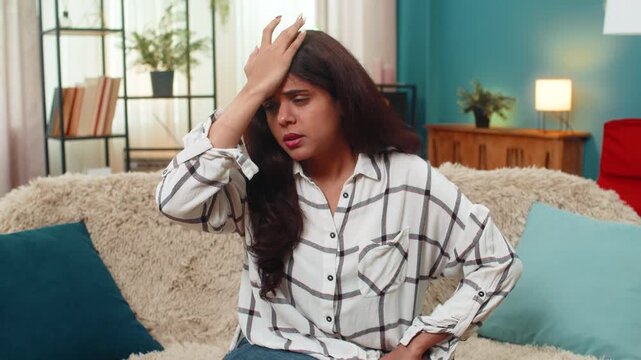 Indian woman at home taps forehead realizing mistake feeling embarrassed deeply and regretful. Arabian girl on sofa makes facepalm gesture showing repentance self blame confusion surprise and anxiety.
