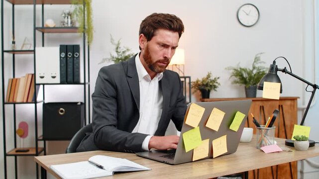 Middle-aged businessman at home office looks at laptop full of stickers for tasks and feels pressure. Freelancer guy at table worries about deadlines types fast and tries to finish plan before end