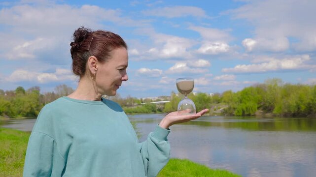 Woman holding hourglass meditating on time passing. Mature woman holding an hourglass while standing by a river. The flowing water and running sand symbolize the relentless passage of time and life