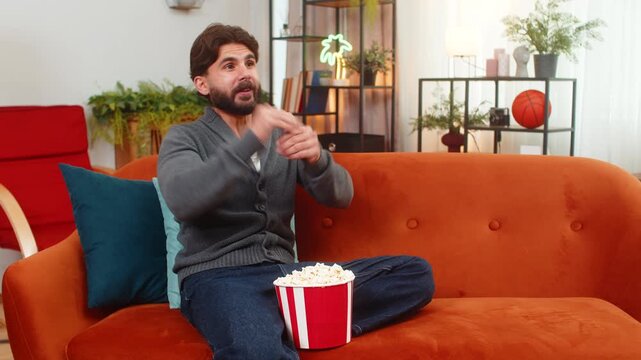 Indian man at home watches series finale on TV holds popcorn tense pause then smiles and claps. Arabian guy on sofa follows last episode feels joy relief and applause after dramatic end on screen