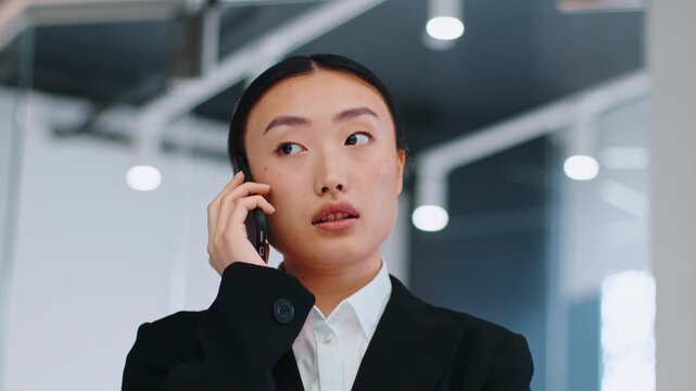 Focused Asian Woman Engaged In Conversation On Smartphone While Standing In Contemporary Office Environment, Wearing Formal Attire. Representing Professional Business Concept.