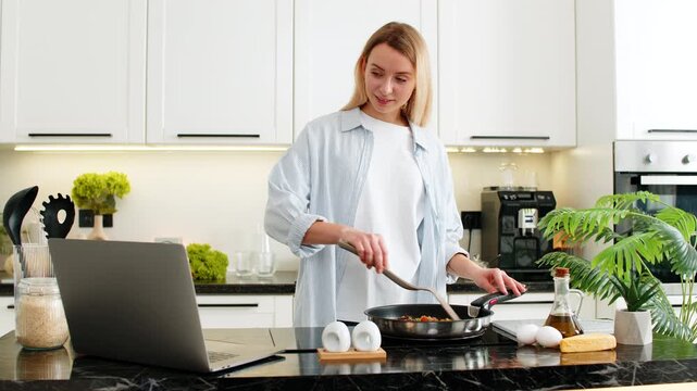 Middle-aged woman fries vegetables in pan on electric cooktop while watching keto recipe on laptop at home kitchen for weight loss goal. Young girl tries low carb lunch prep feeling focused so happy.
