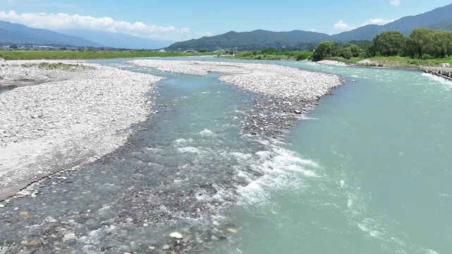 Clear river rushes over smooth stones. Shimmering water carves through rocky bed. Distant mountains rise under bright sky. Vibrant green banks frame flowing current