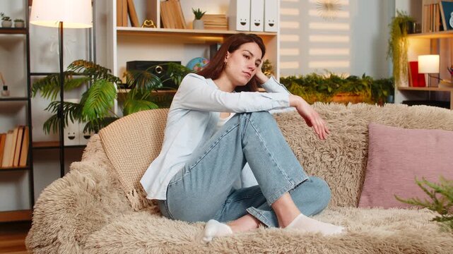 Young woman on home sofa looks sad thinking deeply and sighing over personal worries. Adult girl on home sofa shakes head with worry feeling quite anxious stressed and emotionally overwhelmed indoors.
