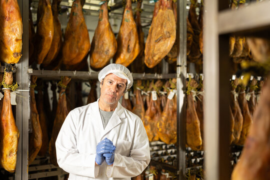 Man employee in white uniform, bathrobe and hat, stands in workshop for production of ham. Working moments, close-knit team, private enterprise