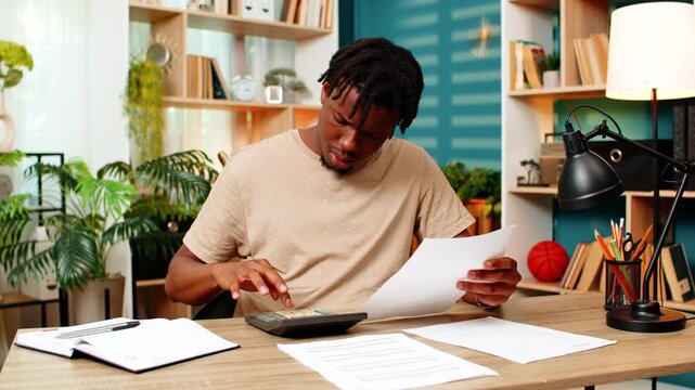 African American young man on home table checks papers and calculator while reviewing budget notes and account records. Black guy on home table checks sums and records for budget control and bills.