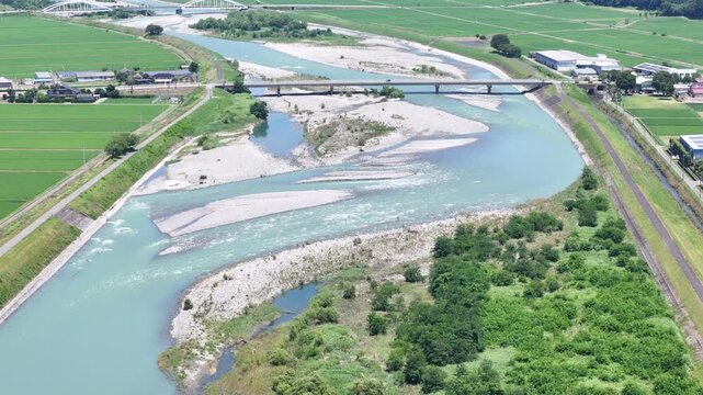 Aerial view of a winding river through green fields. Two bridges span the water, connecting communities. Railway runs parallel, blending with nature