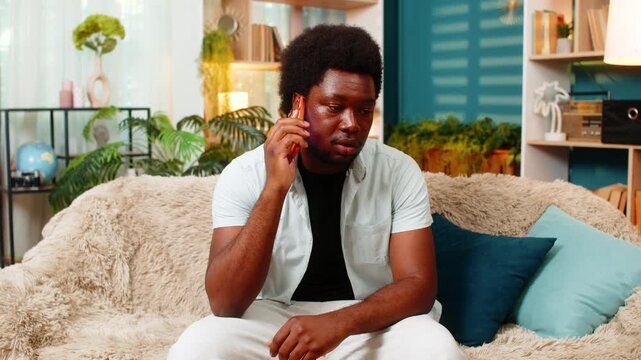 African American young man on home sofa answers smartphone call with calm face in remote talk. Black guy on home sofa speaks by phone for family news support or client call in cozy warm living room.