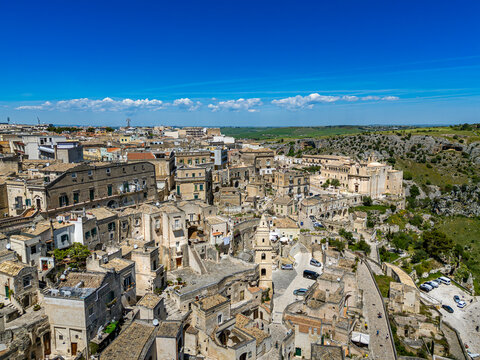 An expansive aerial view overlooking the UNESCO-listed Sassi di Matera, featuring the dense limestone dwellings, the Church of San Pietro Barisano, and the lush green slopes of the Murgia plateau unde