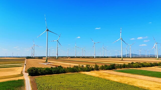 Wind power generators on vast fields under blue sky. Aerial landscape of electricity producing turbines in yellow rural sector during daytime.