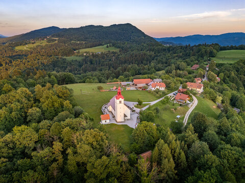 A high-angle landscape view of the hilltop Church of the Holy Trinity (Cerkev svete Trojice) in Sveta Trojica above Moravče, Slovenia, with a brilliant golden sunset illuminating the distant mountains